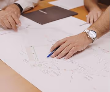 Two people reviewing architectural blueprints on a table, one pointing with a pen.