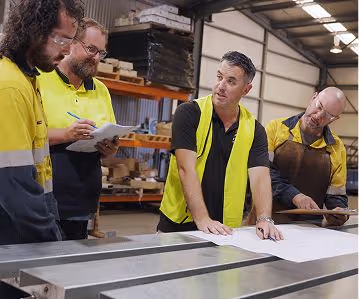 Four men in a workshop with safety vests discuss and review large blueprints on a metal table.