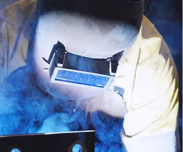 Welder wearing protective mask and yellow sleeves welding metal with blue sparks and smoke.