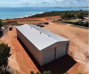 Large metal warehouse with two garage doors sits on red dirt near a coastline with blue ocean and sky.