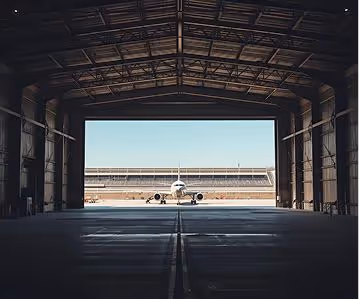 Airplane parked outside viewed through the dark interior of a large hangar.