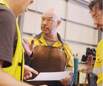 Three workers in safety vests and goggles discuss plans, one holding a paper in an industrial setting.