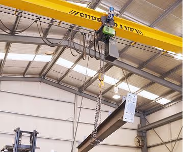 Yellow overhead crane lifting large steel beam inside warehouse with metal roof and walls.