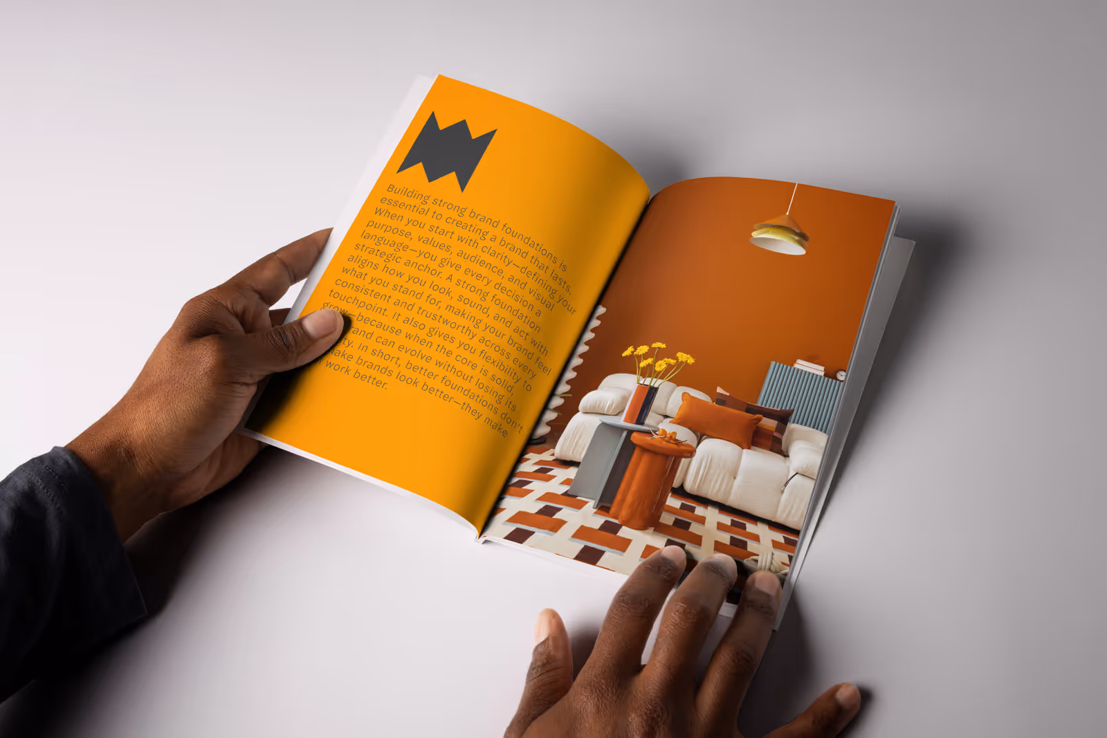 Person holding open a magazine showing a page with text on an orange background and a photo of a modern living room with a white sofa, orange wall, patterned rug, and yellow flowers.