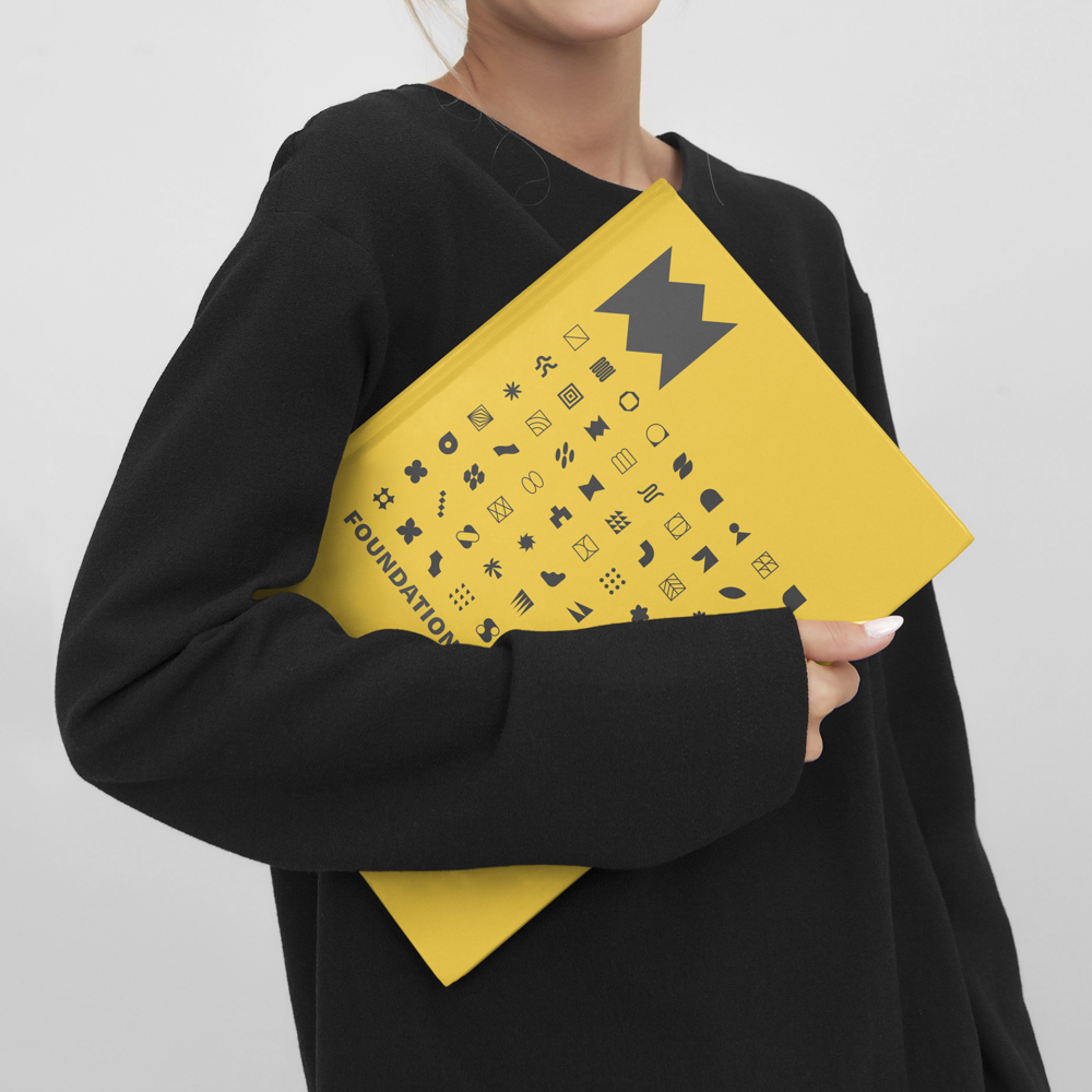 Person in black long-sleeve shirt holding a yellow book titled 'FOUNDATIONS' with black geometric icons on the cover.