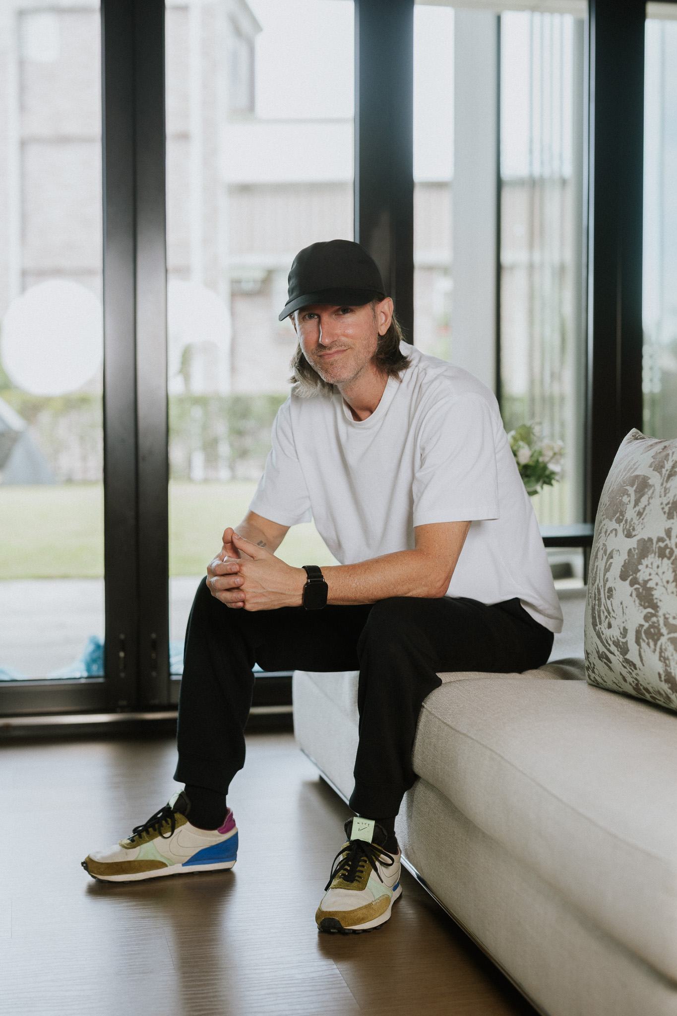 Man in a black cap, white t-shirt, black pants, and colorful sneakers sitting on a beige couch indoors.