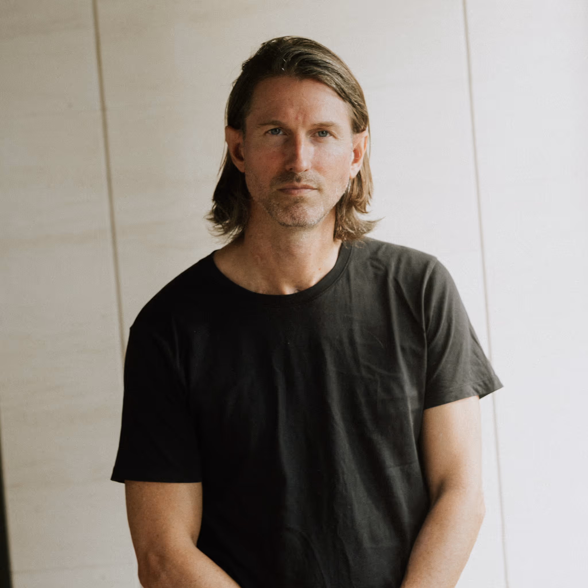 Man with medium-length brown hair wearing a black t-shirt against a beige and white background.