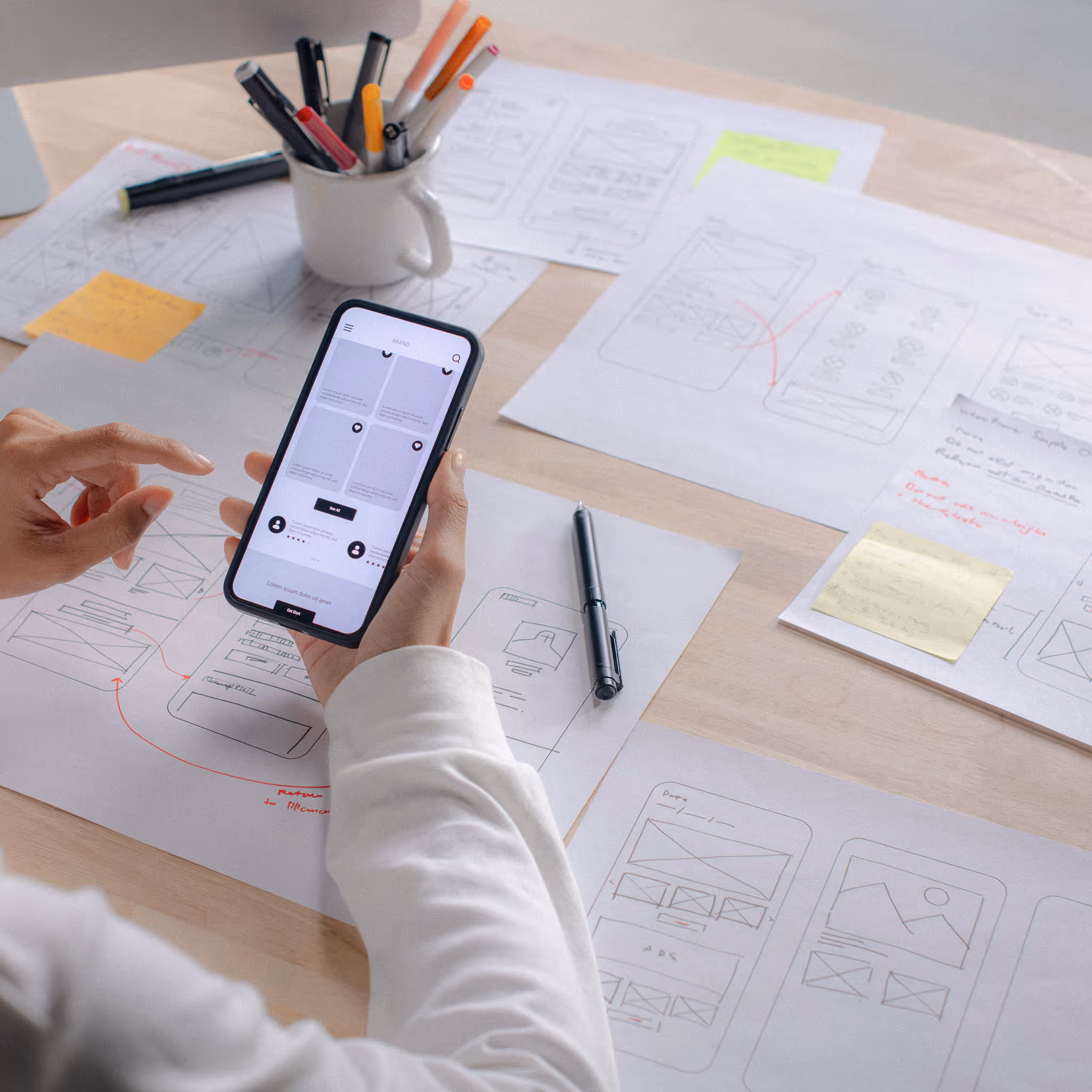 Person holding a smartphone showing a wireframe app screen over a desk covered with hand-drawn UI sketches and colored markers.