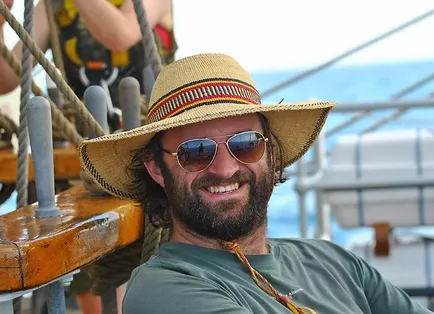 Man with beard wearing sunglasses and a wide-brim straw hat sitting on a boat with wooden railings and ropes.