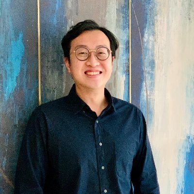 Smiling young man wearing round glasses and a dark button-up shirt standing in front of a blue and beige textured wall.