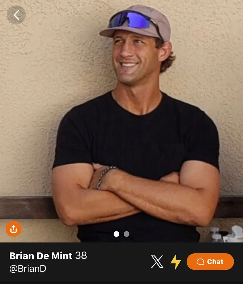 Smiling man wearing a beige cap with reflective sunglasses on top, a black t-shirt, arms crossed, sitting against a textured beige wall.