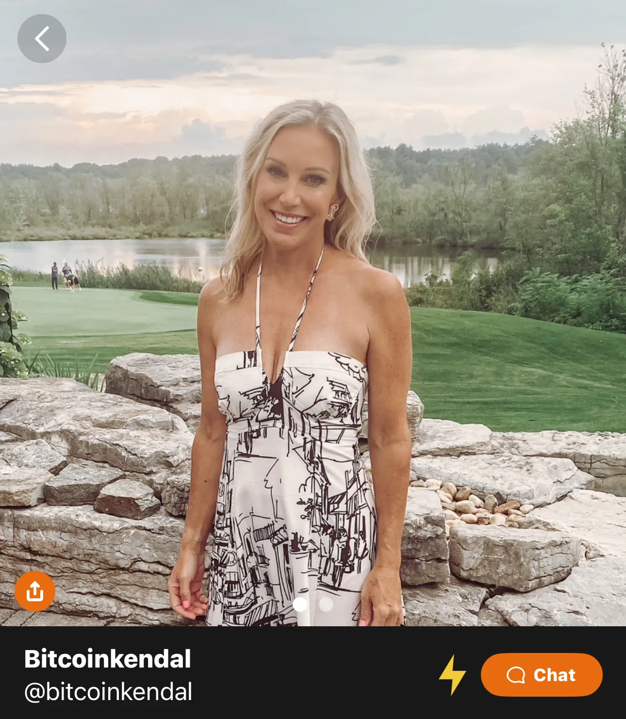 Smiling woman with blonde hair wearing a strapless patterned dress standing in front of stone wall with a lake and trees in the background.