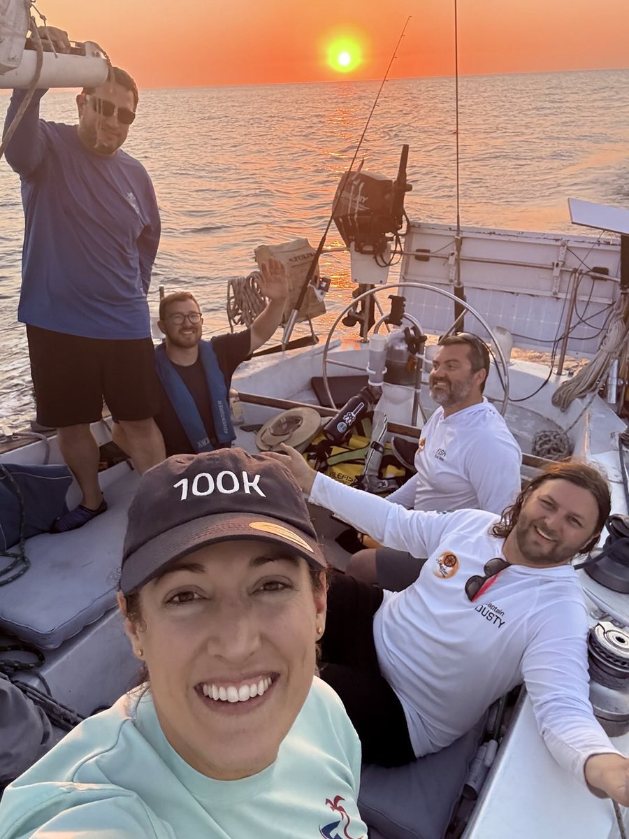 Group of five people smiling and relaxing on a boat during a vibrant sunset over calm sea waters.