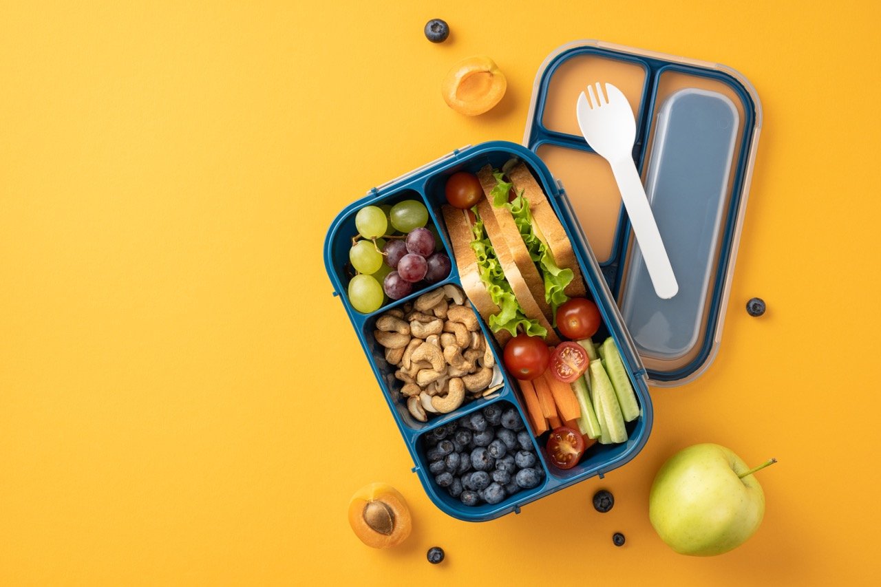 Blue lunchbox with sandwich, grapes, cashews, blueberries, cherry tomatoes, carrot sticks, and cucumber slices on yellow background.