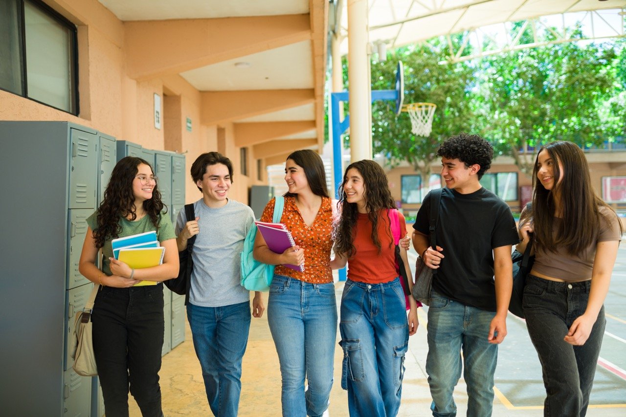 Group of six high school students walking and talking in a school hallway with lockers and a basketball hoop in the background.