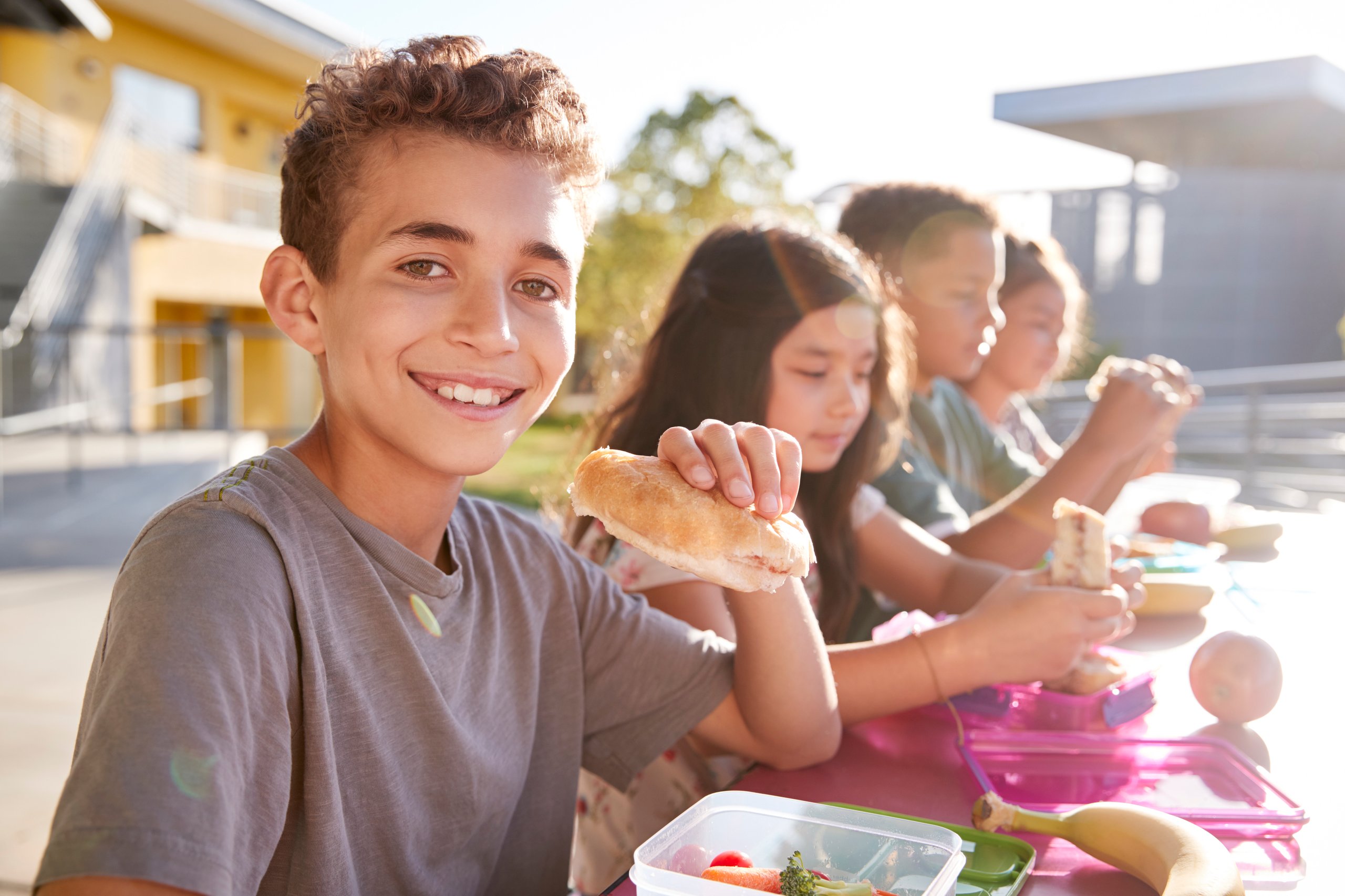 Smiling boy holding a sandwich while sitting at an outdoor table with other children eating lunch.