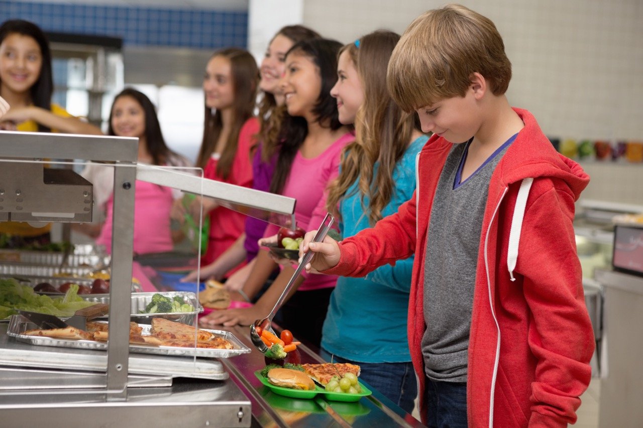 Boy in red hoodie serving vegetables on a tray in a school lunch line with other children waiting behind him.