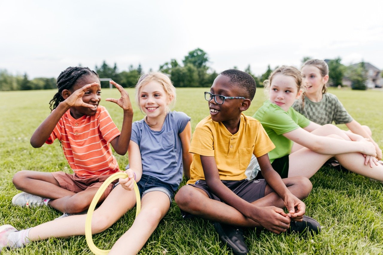 Five children sitting on grass in a park, smiling and interacting, one holding a yellow ring.