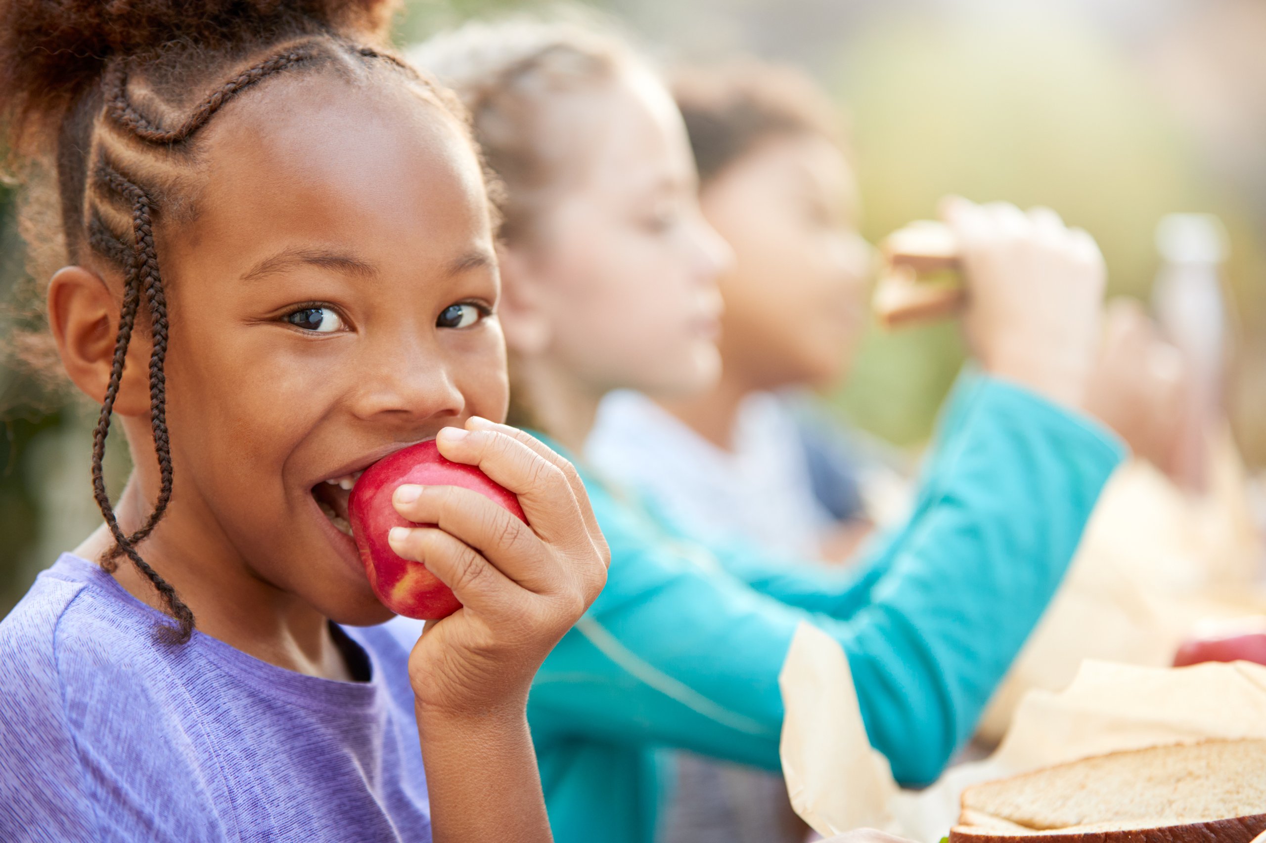 Child with braided hair in a purple shirt biting a red apple, with other children eating sandwiches blurred in the background.
