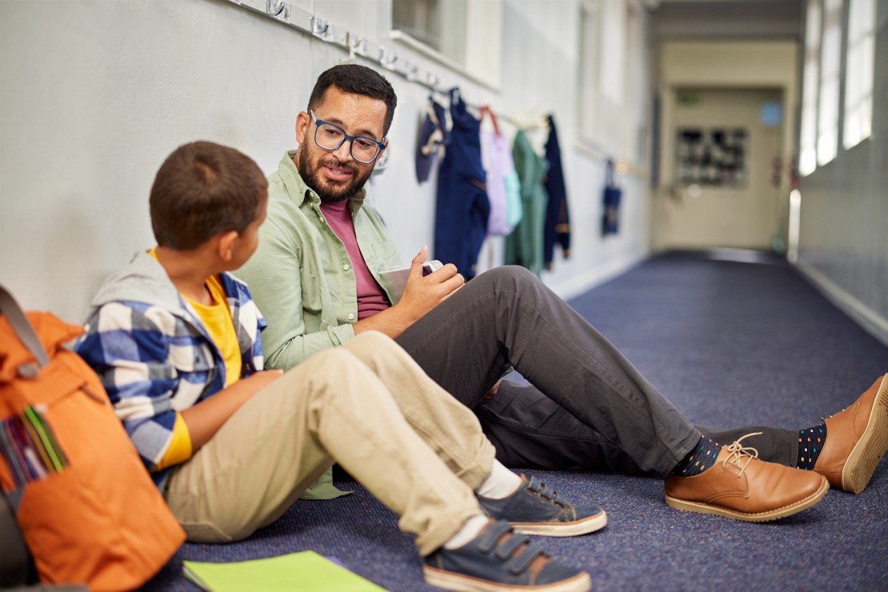 Teacher sitting on the floor in a school hallway talking to a young student.