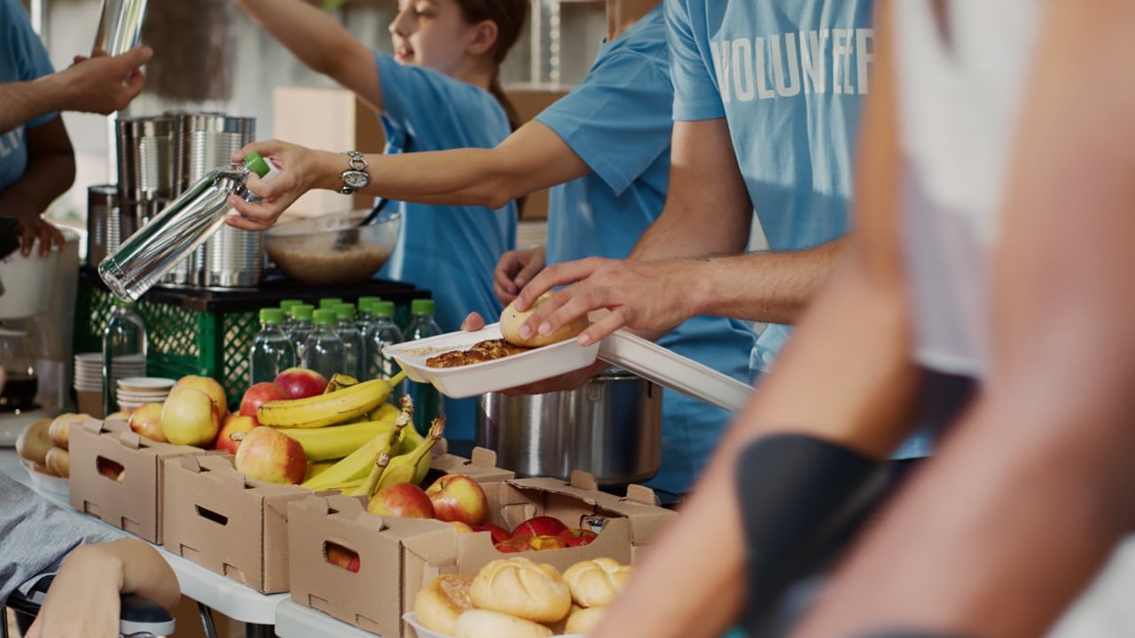 Volunteers in blue shirts serving food, including apples, bananas, bread, and bottled water, at a community food distribution event.