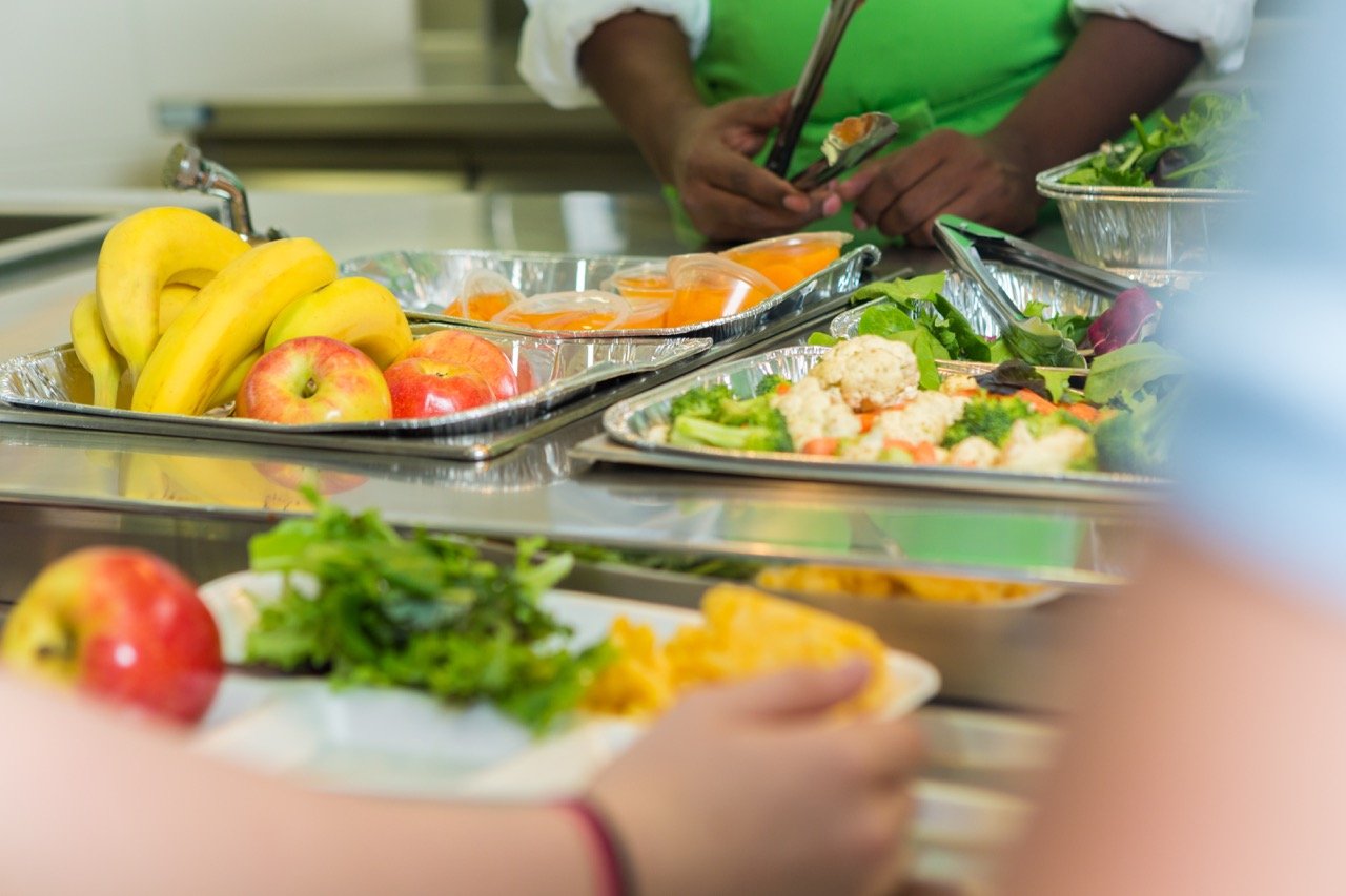 Person serving fresh fruits and vegetables on trays in a cafeteria line.