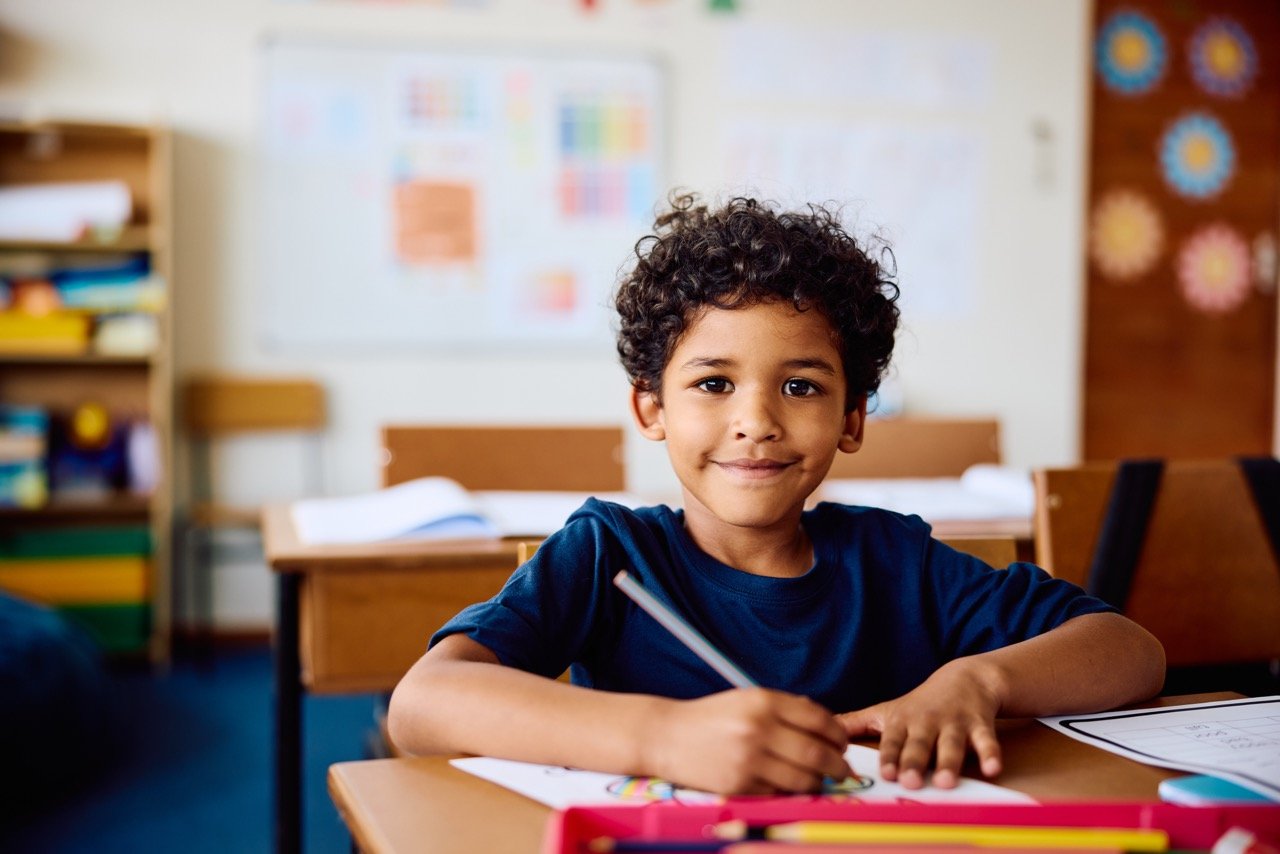 Young boy with curly hair smiling and coloring at a desk in a classroom.