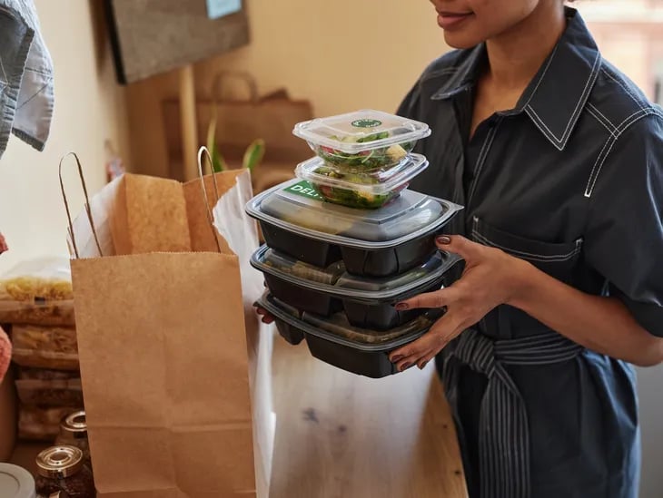 Woman holding a stack of meal prep containers next to a brown paper grocery bag in a kitchen.