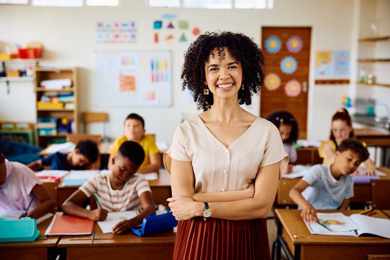Smiling female teacher standing with arms crossed in a classroom with children working at desks.