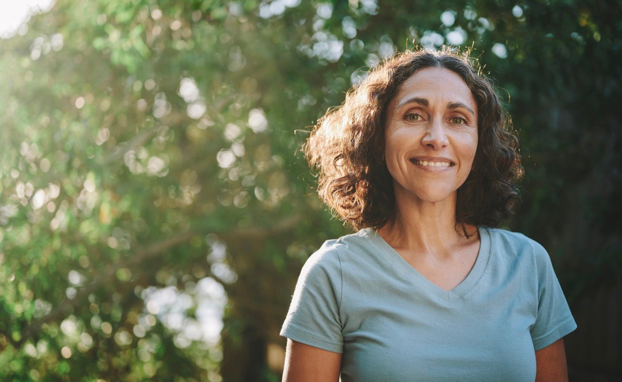 Smiling woman with curly hair wearing a light blue shirt standing outdoors in front of blurred green foliage.