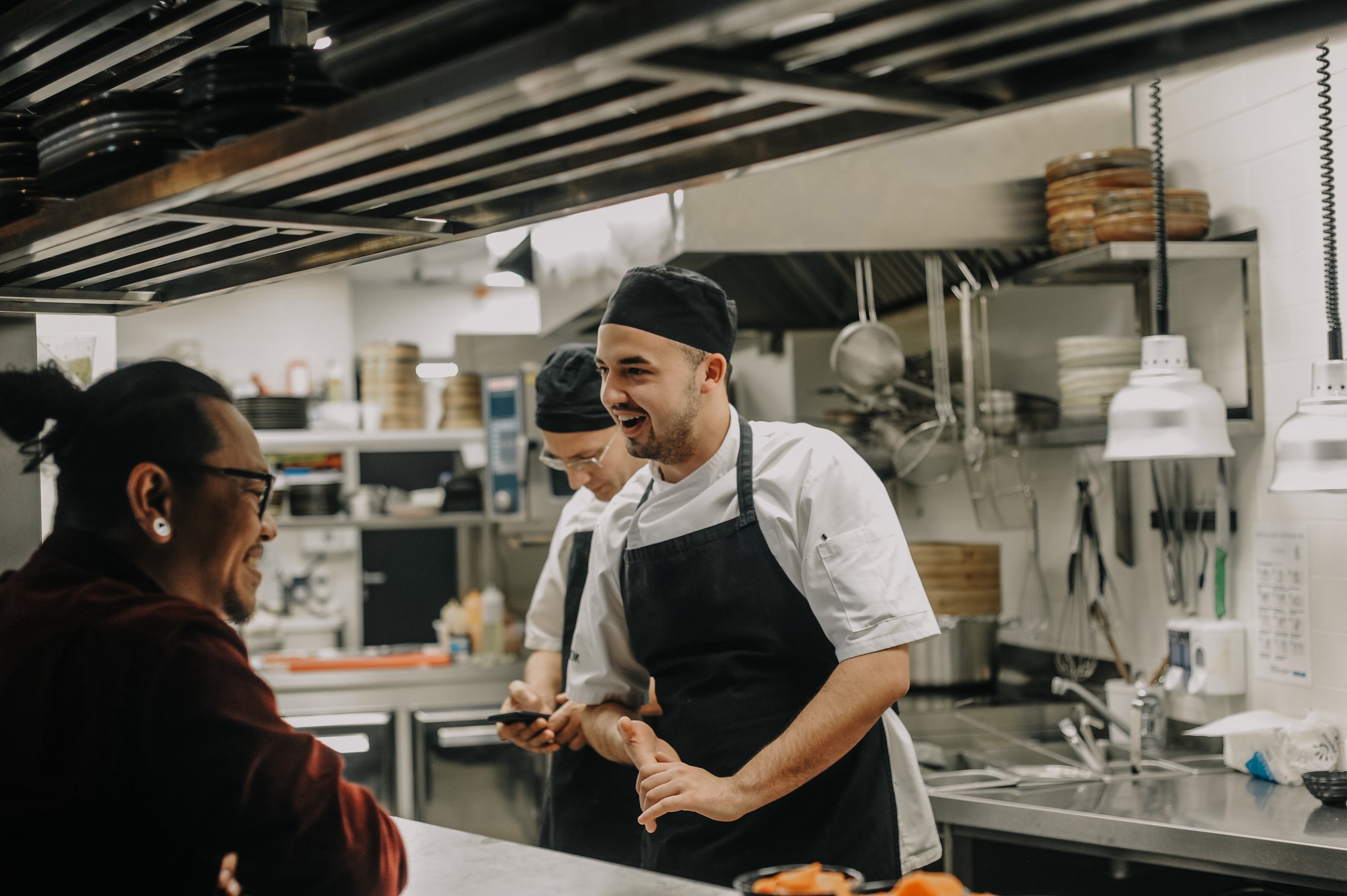 Two chefs in black aprons and hats smiling and talking to a customer at a restaurant kitchen counter.