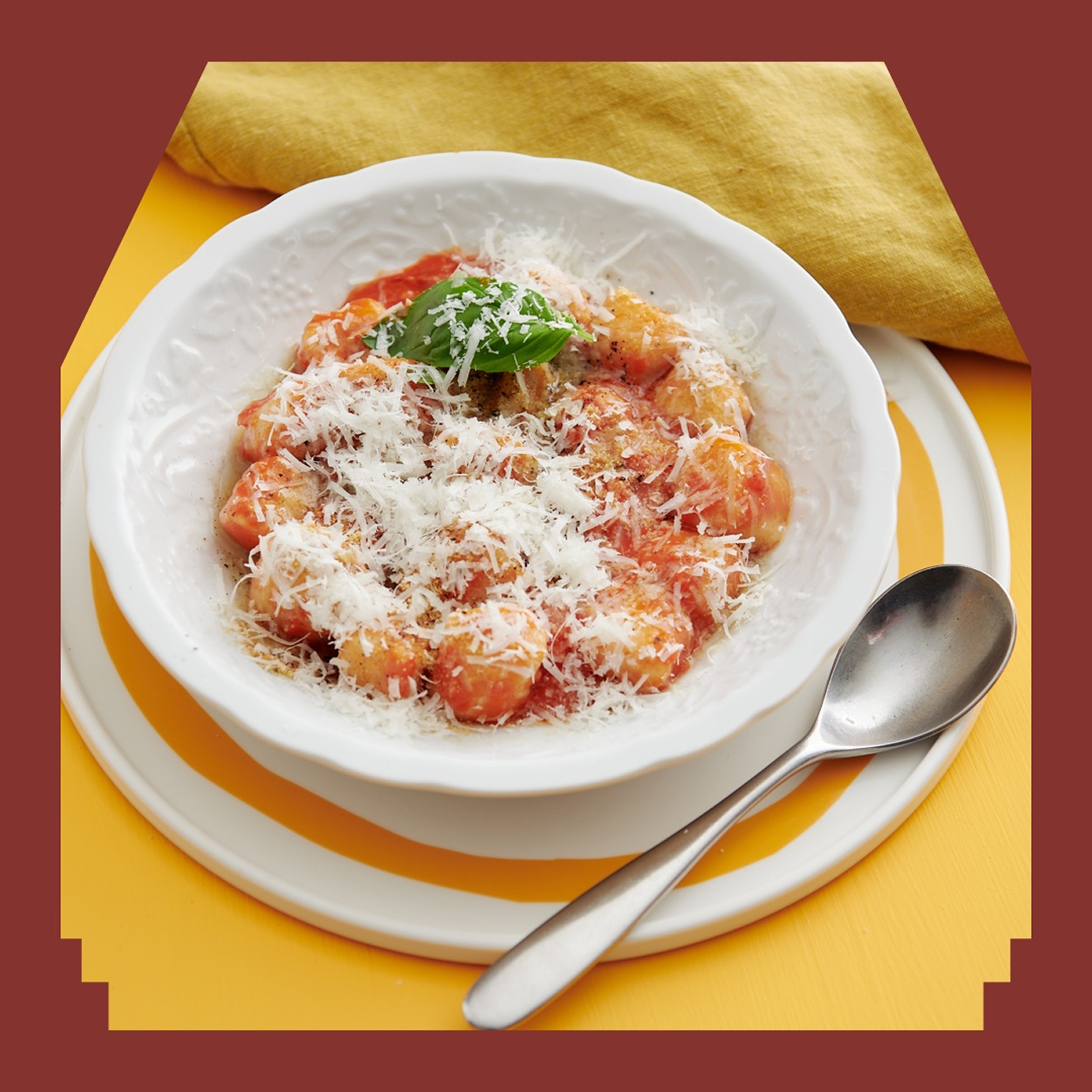 Bowl of pasta with tomato sauce and shredded cheese on top, sitting on a yellow and white placemat and yellow tablecloth, featured in a brown frame.