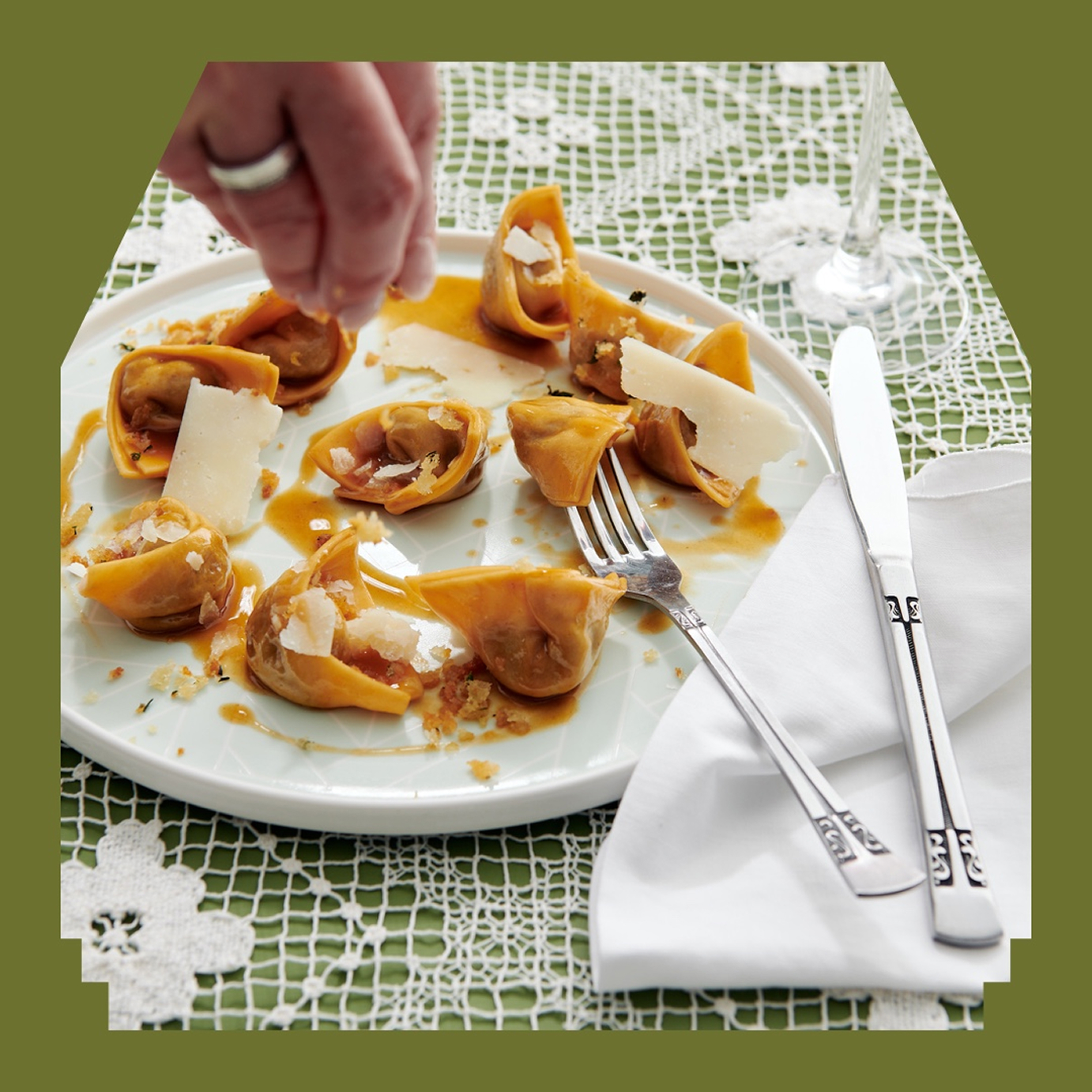 Hand sprinkling cheese onto pasta on a white plate, sitting on a tablecloth with an olive green border.