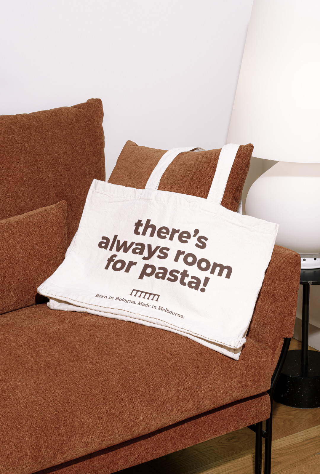 White tote bag with the text "there’s always room for pasta!" resting on a brown fabric sofa next to a white lamp.