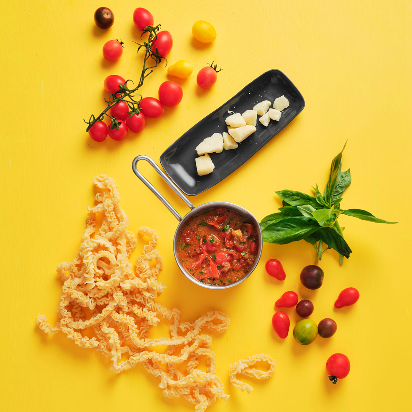 Top down mise en place of pasta, sauce, cherry tomatoes, cheese and basil, scattered on a bright yellow surface.