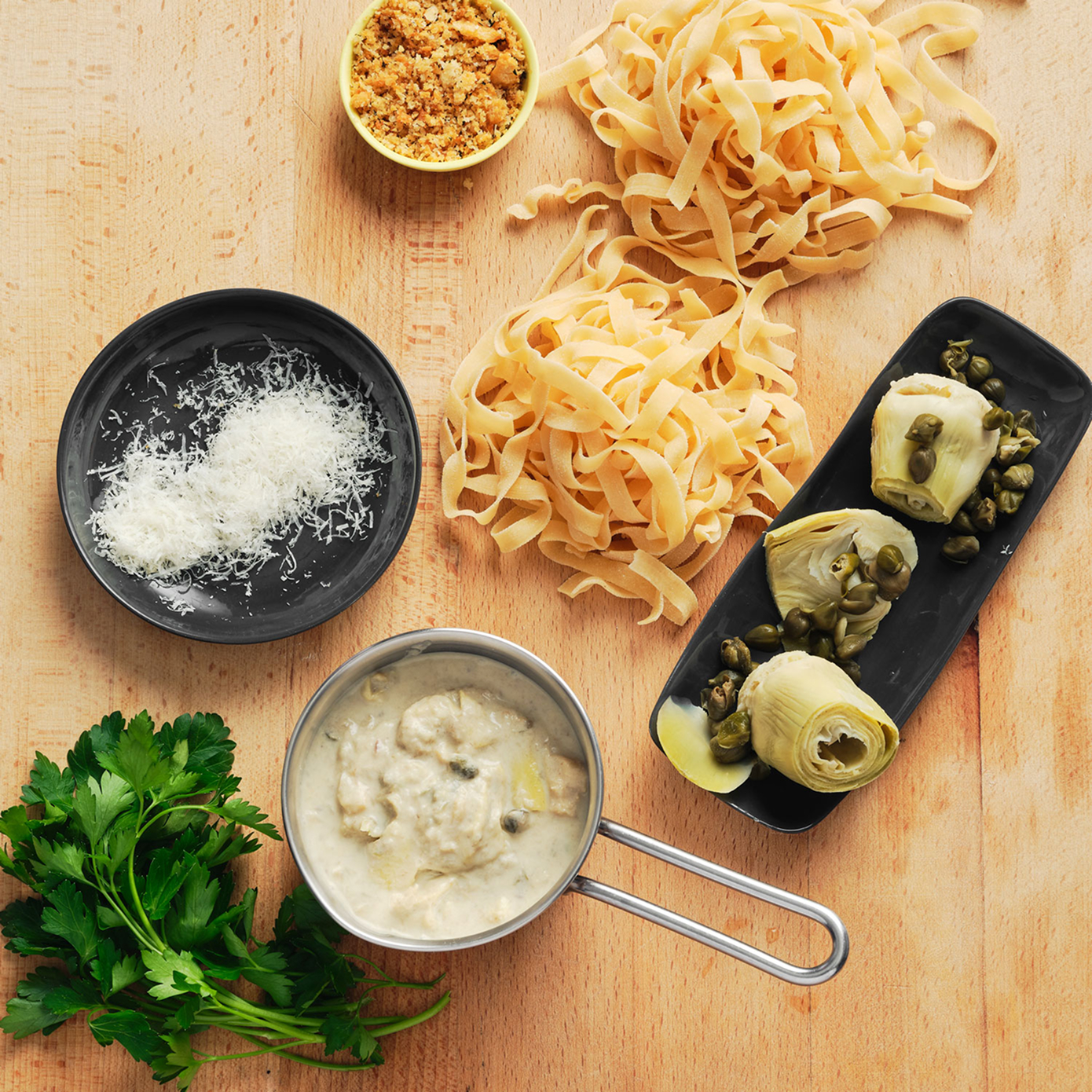 Top down mise en place of pasta, a cream sauce, artichoke hearts, cheese and pangrattato, scattered on a wooden surface.