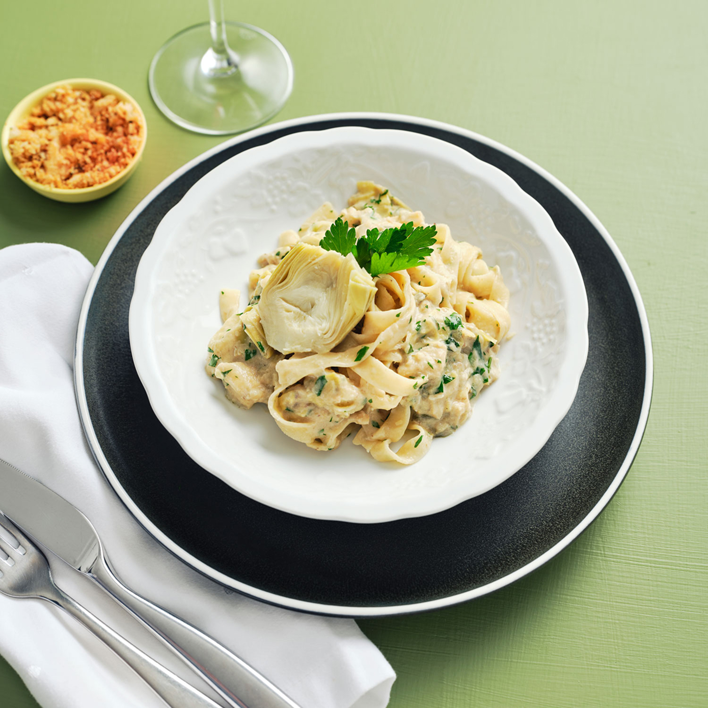 Bowl of creamy fettuccine pasta sitting on a black plate, on a green surface with pangrattato and a wine glass nearby.