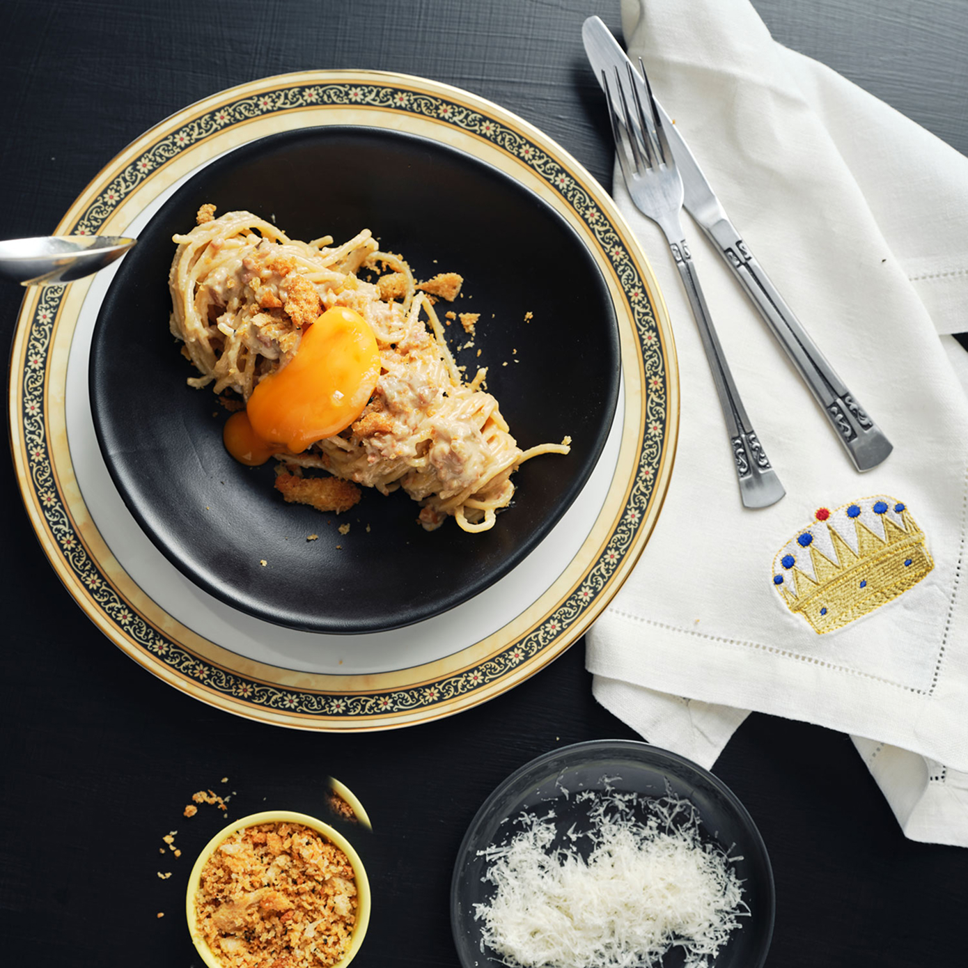 Bowl of spaghetti carbonara with an egg yolk on top, sitting on a black table with pangrattato, cheese, and an embroidered napkin nearby.