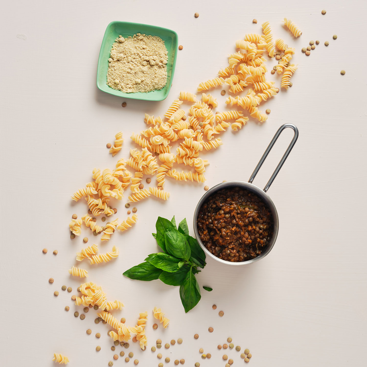 Mise en place of lentil fusilli scattered amongst some basil leaves, a lentil sauce and some powdered cheese, all on a light cream surface.