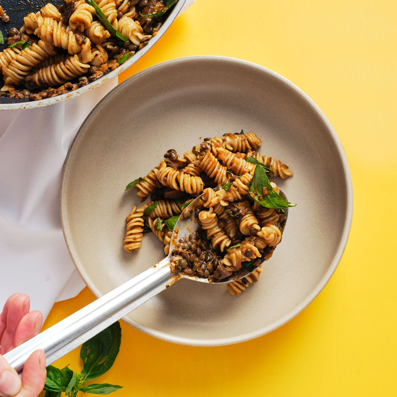 Large metal spoon scooping lentil fusilli from a cream bowl, sitting on a bright yellow surface.