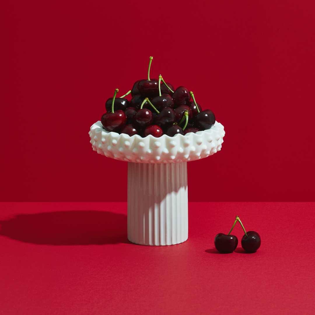 White pedestal with cherries piled on top of it, in front of a red background.