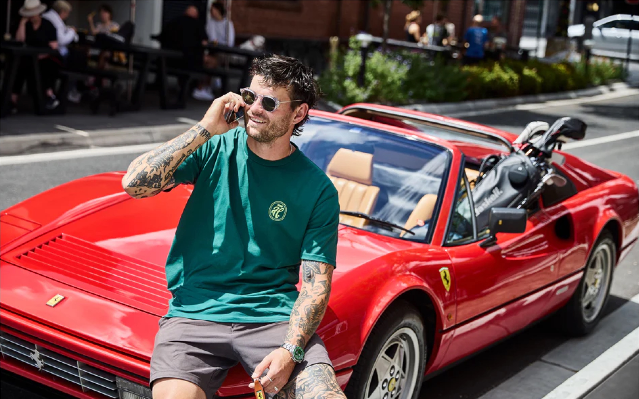 Man wearing a green Haskell shirt, talking on a mobile phone and sitting on the hood of a red car with a golf bag sitting in it.