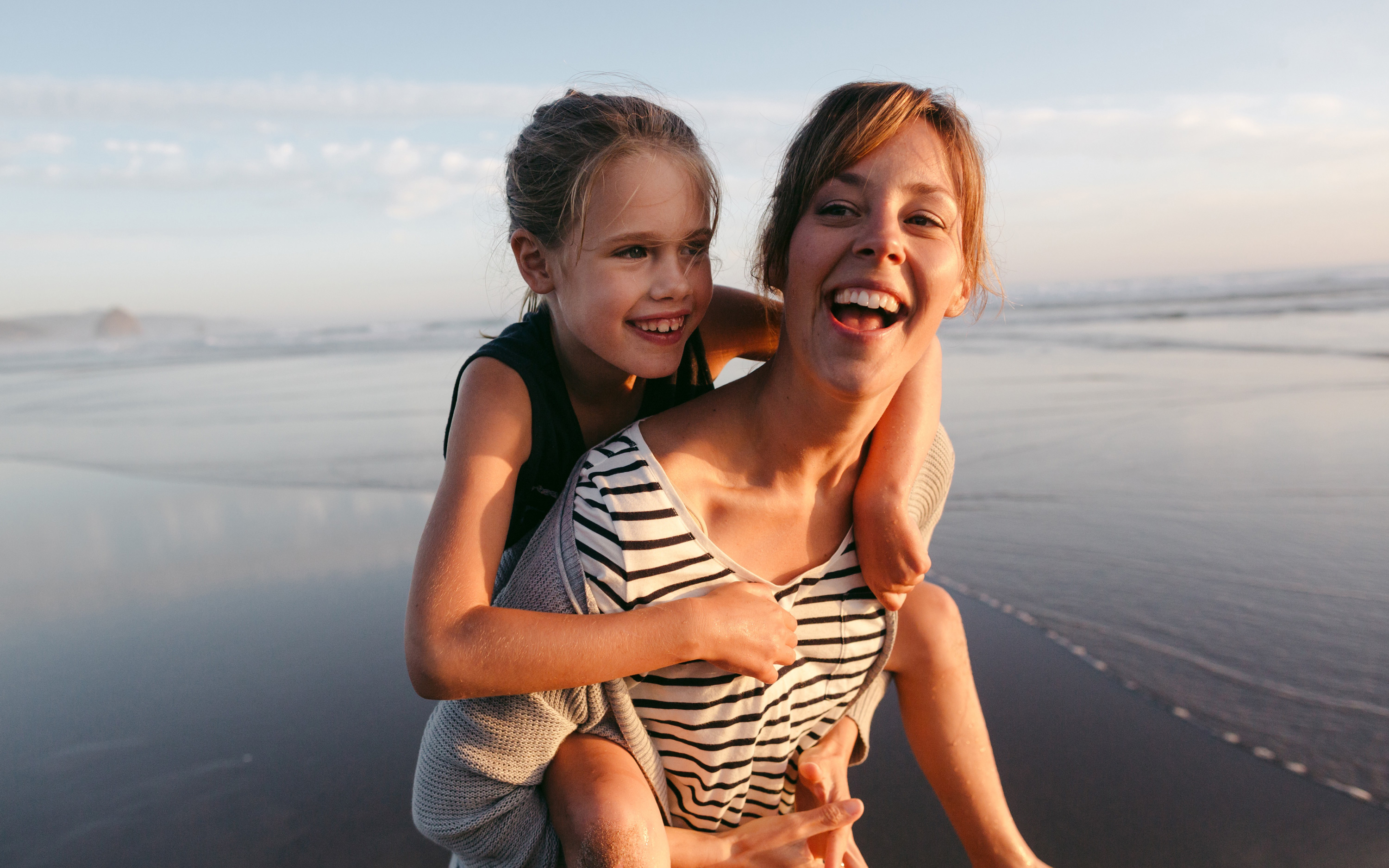 Mother giving daughter a piggy back ride while walking along the water's edge at the beach.