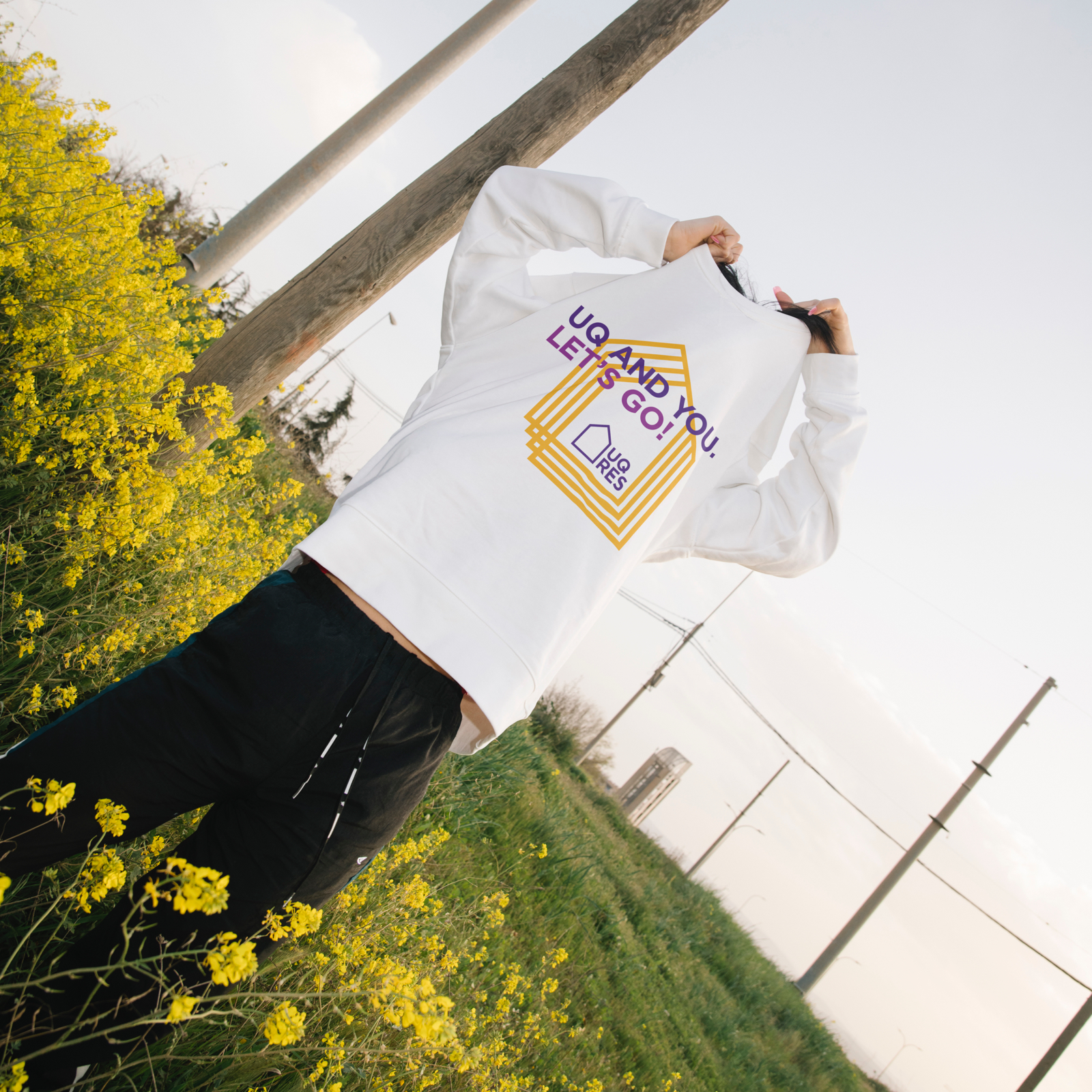 Person standing in a field of yellow flowers, pulling off a white sweatshirt with a graphic of yellow UQ Res house logos and text which reads "UQ and You. Let's Go!".