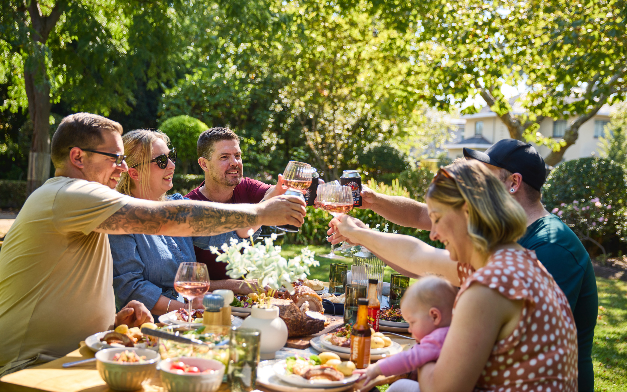 Group of men and women cheersing their drinks over a table setting, in a sunny garden.