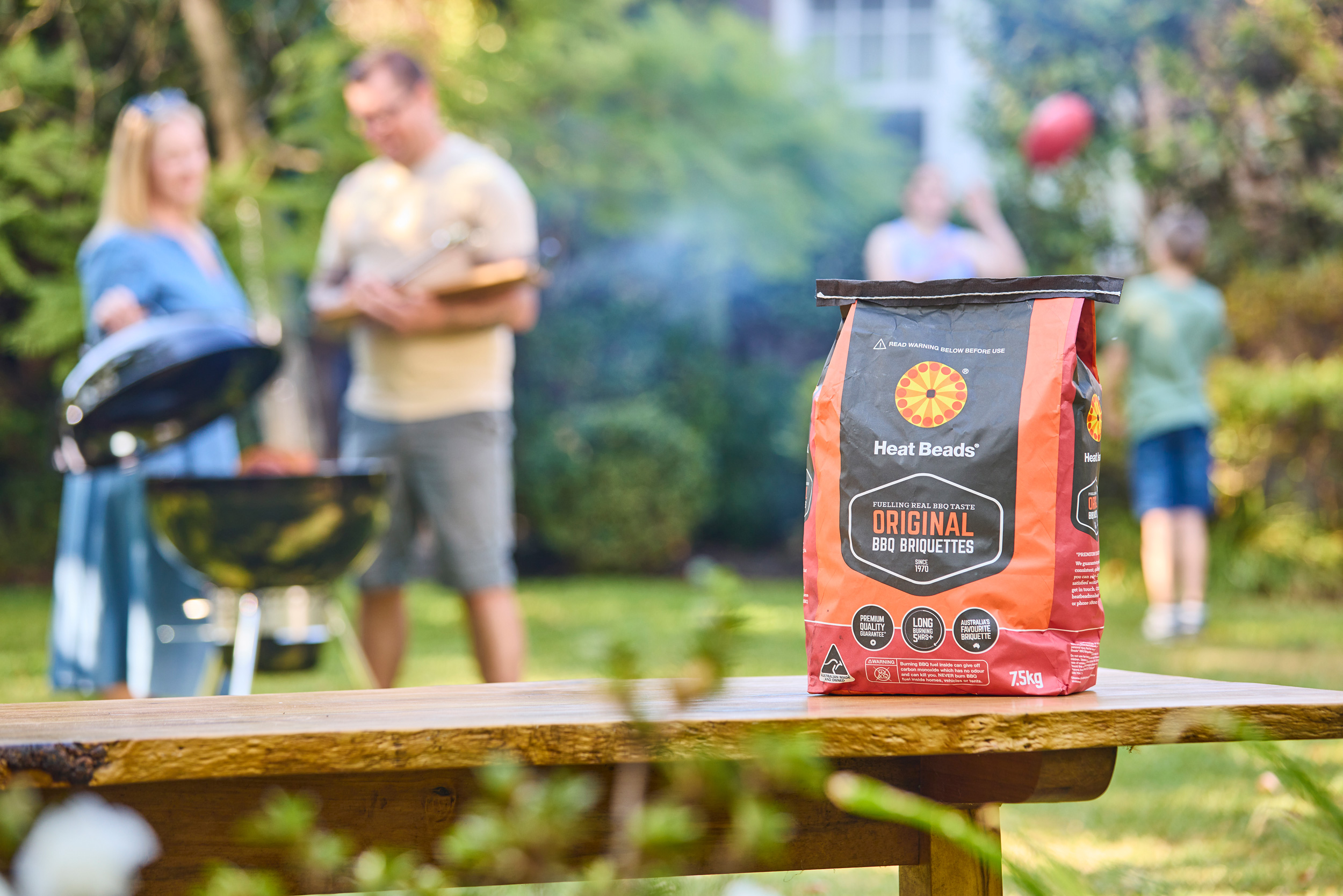 Heat Beads briquettes bag on a table, with family cooking a barbecue out of focus in the background.