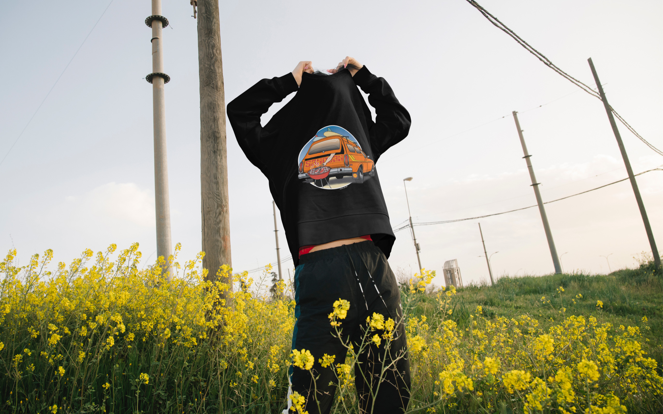 Person standing in a field of flowers, pulling off black sweatshirt with illustration of an orange Holden Sandman car on it.