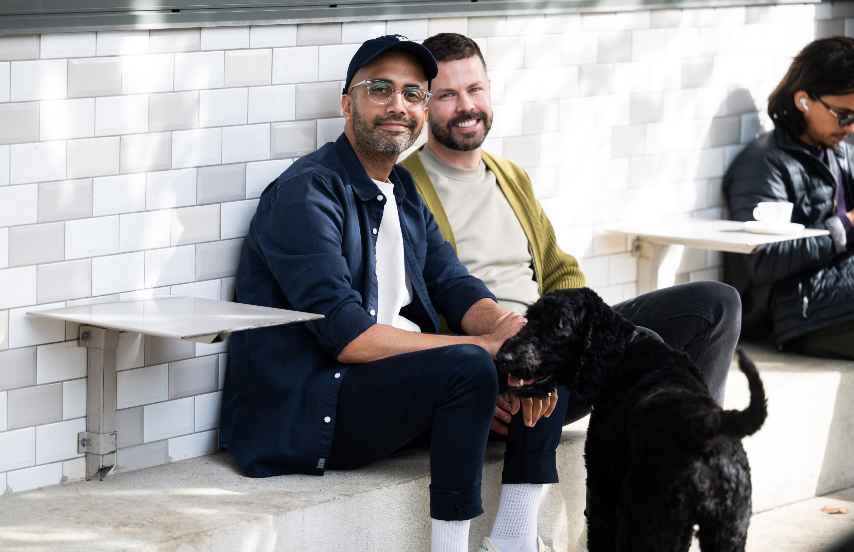 Two men sitting side-by-side in front of a grey and white tiled wall, smiling at the camera and joined by a black dog. 
