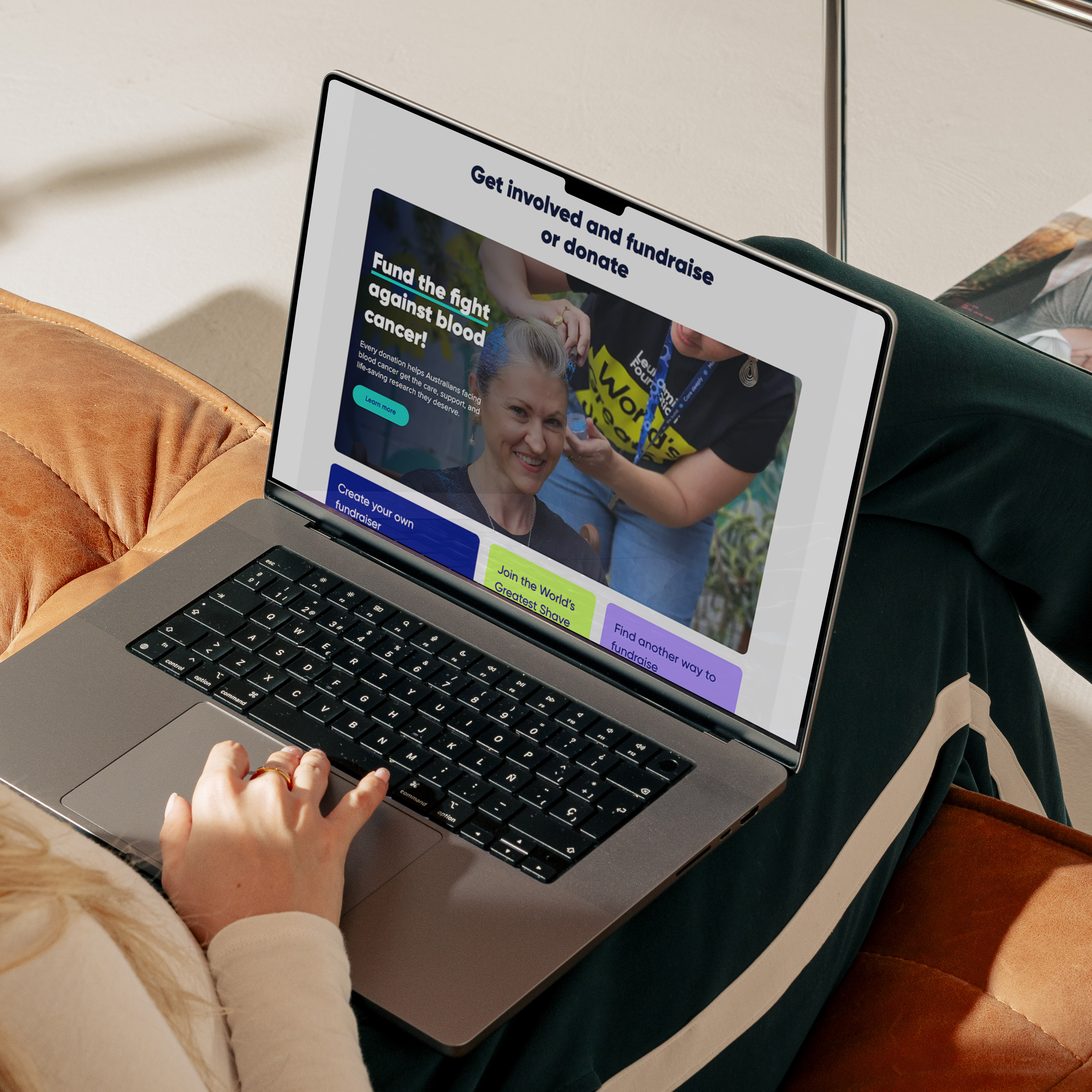 The Leukaemia Foundation website shown on an open laptop, being used by a woman with green tracksuit pants sitting on an orange couch.