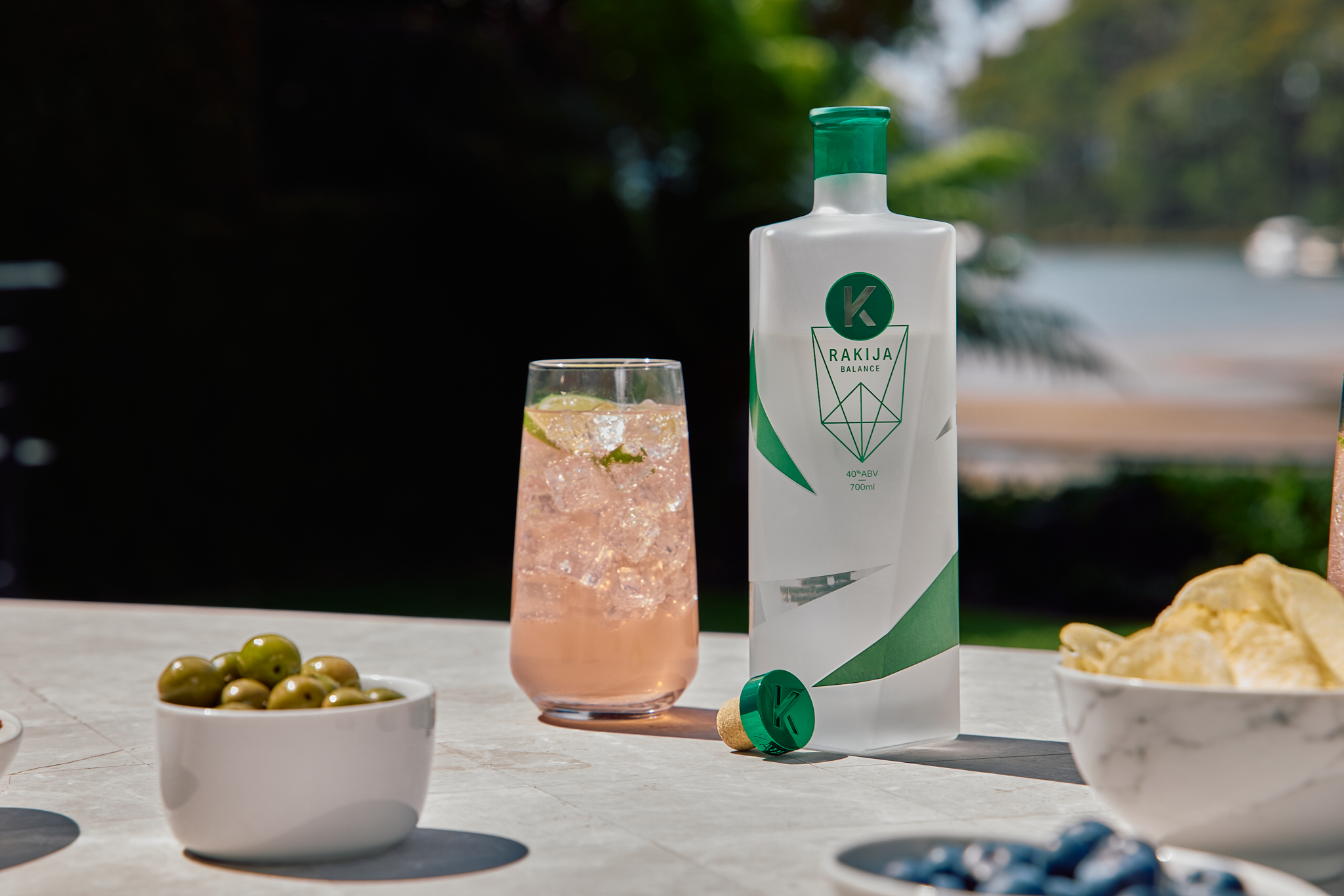 Frosted Kabina bottle of Rakija sitting on a table next to a peach-coloured mixed drink, with small bowls of olives and chips nearby.